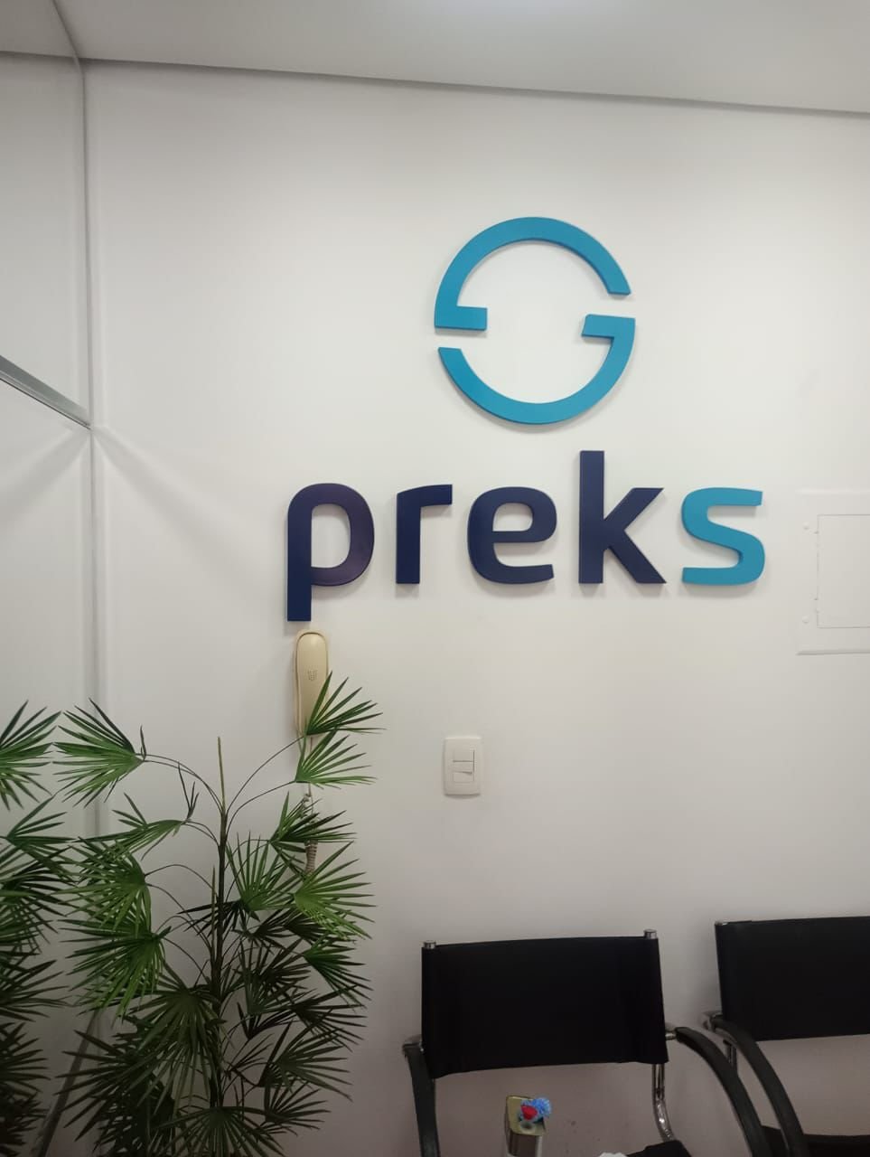 Logo e nome da Preks aplicados em parede interna com letras azuis e símbolo circular.