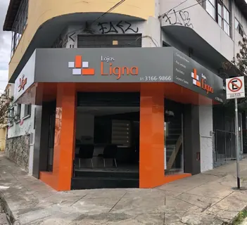 Miniatura: Fachada de esquina da Lojas Ligna com painéis cinza e colunas laranja em perspectiva urbana.