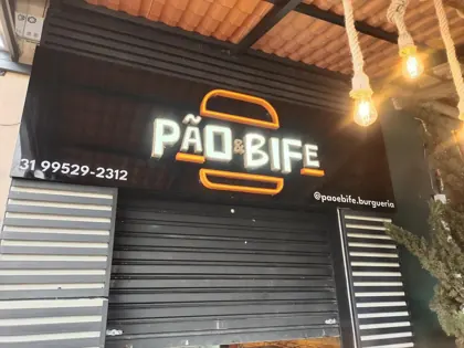 Miniatura: Fachada da Pão & Bife com letreiro neon branco e laranja vista com parte do interior aparente.